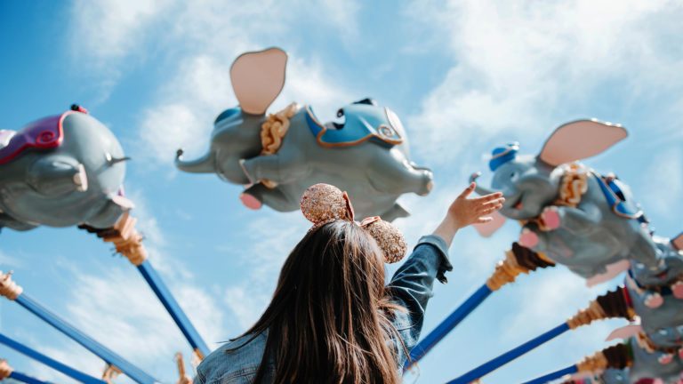 Woman at Walt Disney World wearing Minnie Mouse ears waves at the Dumbo the Flying Elephant ride in the Magic Kingdom.