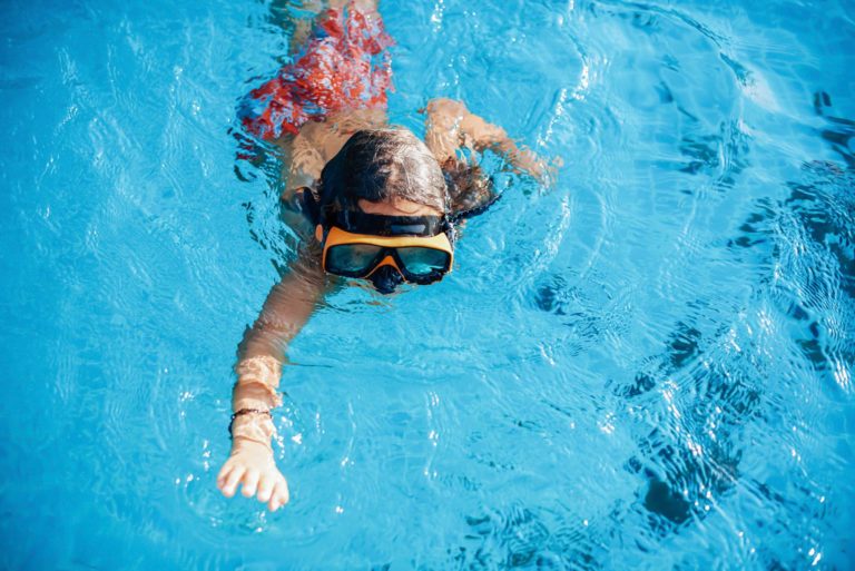 Active little boy swims in a pool in swimsuit with goggles.