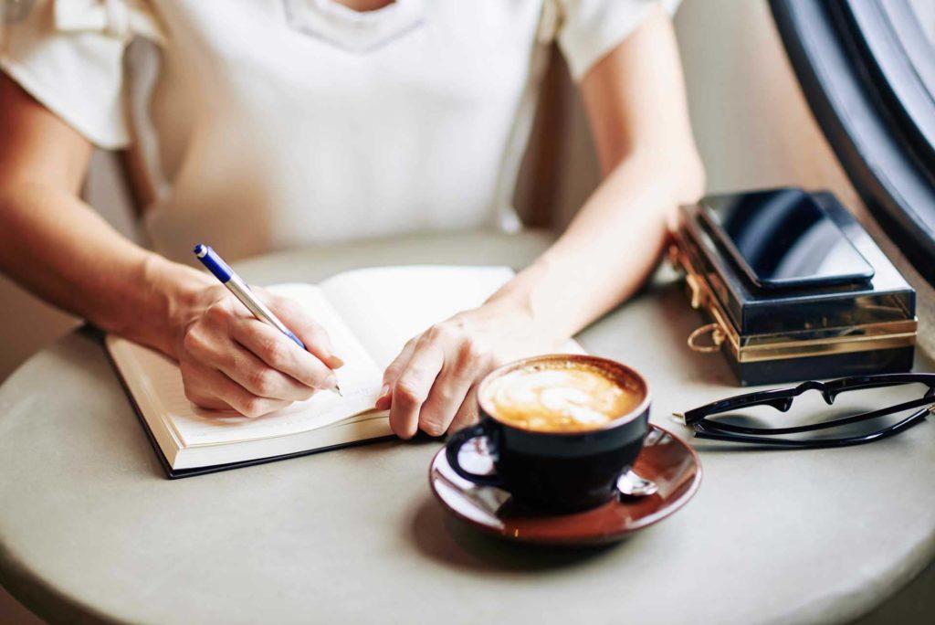 Woman writing in a journal in a coffee shop while drinking a latte