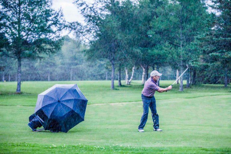 Hombre jugando golf bajo la lluvia mientras cubre sus palos de golf con un paraguas