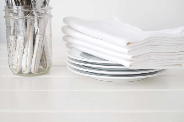 Conjunto de platos blancos y servilletas de lino apiladas junto a un frasco de utensilios