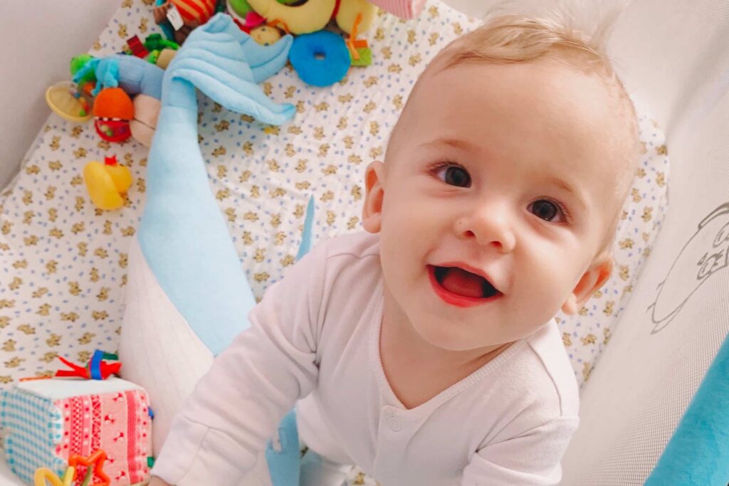Happy baby boy surrounded by toys in a pack 'n play crib