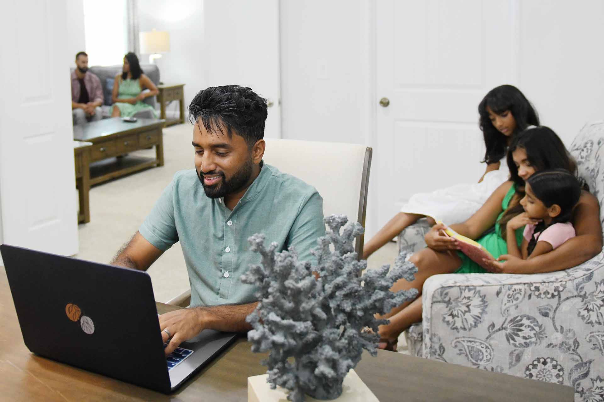 Father working on a laptop while his family reading a book behind him